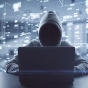 Ein mit einer grauen Hoodie bekleideter Hacker, dessen Gesicht im Schatten der Kapuze nicht erkannbar ist, sitzt vor einem grauen Laptop. Im Hintergrund sind elektronische Icons, die den Cyberspace darstellen sollen. Es geht um Cybersicherheit.