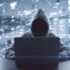 Ein mit einer grauen Hoodie bekleideter Hacker, dessen Gesicht im Schatten der Kapuze nicht erkannbar ist, sitzt vor einem grauen Laptop. Im Hintergrund sind elektronische Icons, die den Cyberspace darstellen sollen. Es geht um Cybersicherheit.