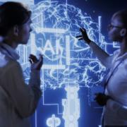 Ein junger Mann und eine junge Frau stehen vor einer dunkelblauen, von hinten mit Licht bestrahlten Wand, auf der KI auf dem HIntergrund eines Gehirns steht.