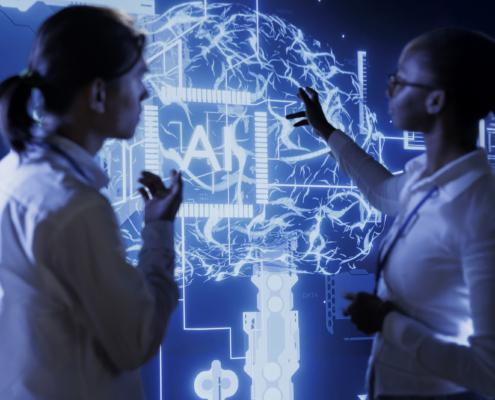 Ein junger Mann und eine junge Frau stehen vor einer dunkelblauen, von hinten mit Licht bestrahlten Wand, auf der KI auf dem HIntergrund eines Gehirns steht.